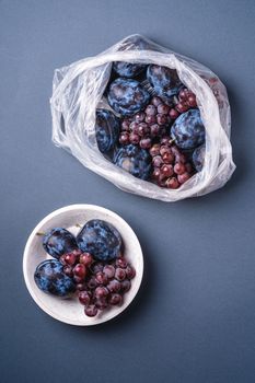 Fresh ripe plum fruits and grape berries in plastic bag package and in wooden bowl on minimal blue grey background, top view