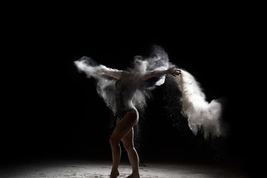 beautiful caucasian woman in a black bodysuit with a sports figure is dancing in a white cloud of flour on a black background