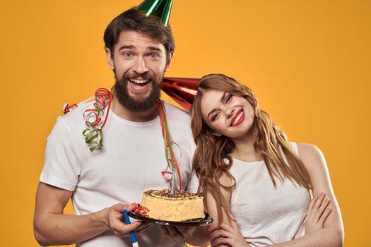 Man and woman birthday festive cake yellow background and caps on the headv. High quality photo