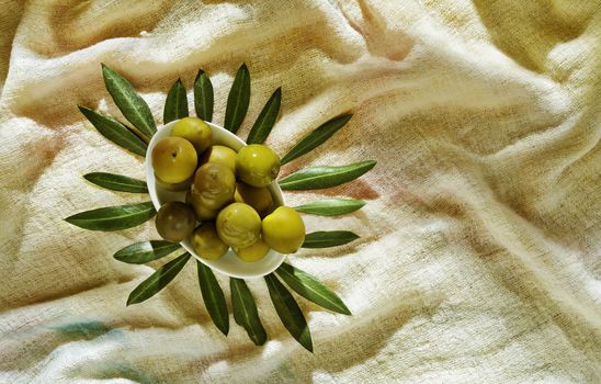 Group of green olives ina white bowl on colored cotton cloth , green leaves surround the bowl