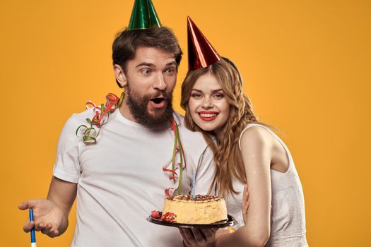 Man and woman birthday festive cake yellow background and caps on the headv. High quality photo