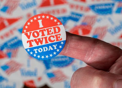Finger with I Voted Twice button in front of many stickers given to US voters in Presidential election to illustrate vote fraud