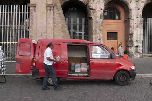 Rome, Italy - June 27, 2010: A man collects maps and guides in a red van, which he has for sale, from a street stall for tourists, next to the Colosseum in Rome.