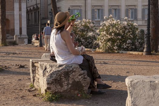 Rome, Italy - June 27, 2010: A tourist woman, wearing a hat, rests sitting on a stone block, drinking a beer, in the area of the Roman Forum, Rome.