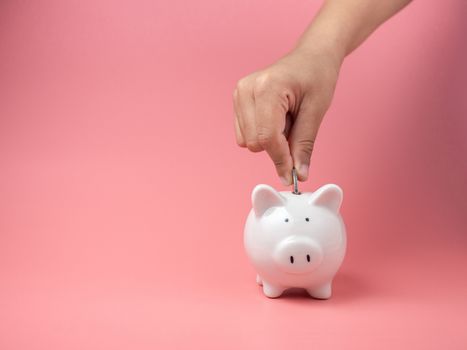 White piggy Bank on a pink background and human hand putting coin in piggy bank. copyspace for design.