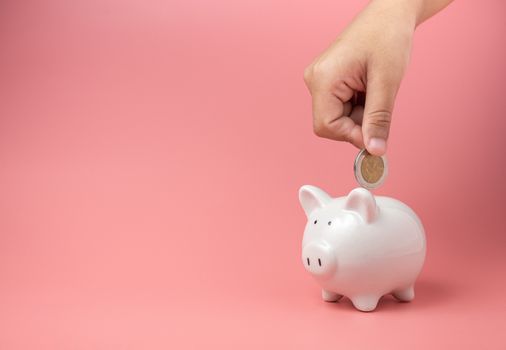 White piggy Bank on a pink background and human hand putting coin in piggy bank. copyspace for design.