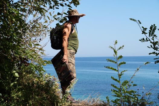 a man with a backpack stands on a high bank and looks at the sea.