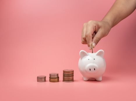 White piggy Bank and coin and human hand putting coin in piggy bank on a pink background. Copy space for design.