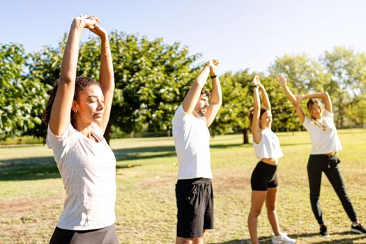 Beautiful young curly hispanic woman teach training with closed eyes together with her three friends in the nature - Cute latina athletics instructor stretching her arms with closed eyes in the park