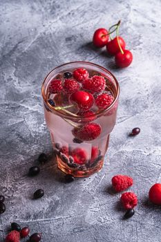 Fresh cold sparkling water drink with cherry, raspberry and currant berries in red faceted glass on stone concrete background, summer diet beverage, angle view