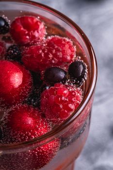 Fresh cold sparkling water drink with cherry, raspberry and currant berries in red faceted glass on stone concrete background, summer diet beverage, angle view macro