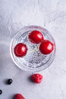 Fresh cold sparkling water drink with cherry, raspberry and currant berries in transparent glass on stone concrete background, summer diet beverage, top view macro