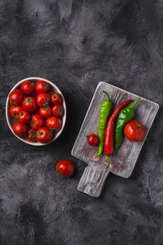 Fresh ripe tomatoes in bowl near to old wooden cutting board with hot chili peppers, dark stone concrete background, top view