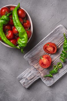 Fresh ripe tomatoes in bowl near to old wooden cutting board with hot chili peppers, stone concrete background, top view