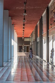 A woman from behind pushes a stroller under the arcades of the city paved with red and white marble, the image of the person is reflected in a shop window