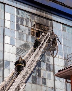 Russia, Kaluga - SEPTEMBER 08, 2020: A team of firefighters in protective suits and helmets on the stairs extinguishing a fire in a building with a broken window and blue roof.