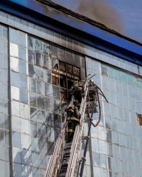 Russia, Kaluga - SEPTEMBER 08, 2020: A team of firefighters in protective suits and helmets on the stairs extinguishing a fire in a building with a broken window and blue roof.