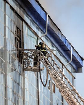 Russia, Kaluga - SEPTEMBER 08, 2020: A team of firefighters in protective suits and helmets on the stairs extinguishing a fire in a building with a broken window and blue roof.