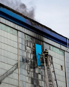 Russia, Kaluga - SEPTEMBER 08, 2020: A team of firefighters in protective suits and helmets on the stairs extinguishing a fire in a building with a broken window and blue roof.