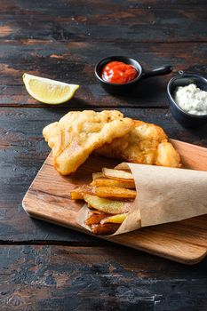 British Traditional Fish and chips with mashed peas, tartar sauce inn crumpled paper cone on wood chopping board dip and lemon - fried cod, french fries, lemon slices, tartar sauce, ketchup tomatoe served in the Pub or Restaurant over old wooden planks dark table side view close up .