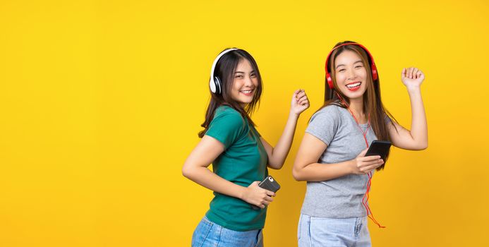 Two Happiness Asian smiling young woman wearing wireless headphones for listening music via smart mobile phone and dancing on isolated yellow color background, Lifestyle and leisure with hobby concept