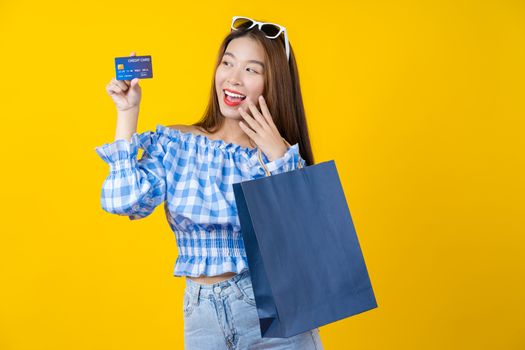 Happy Asian smiling woman using credit card and Carrying a shopping coloful bag for presenting online shopping on isolated yellow color background, copy space and studio, black friday season sale