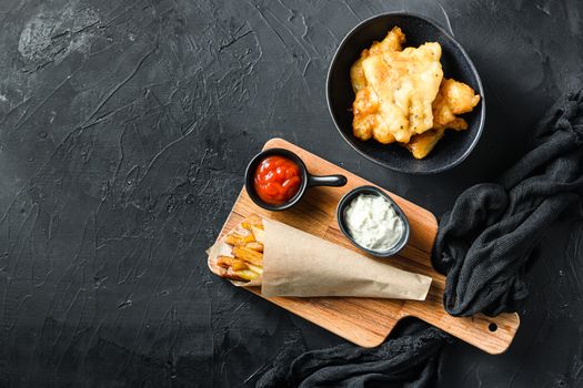 Fish and chips in a paper cone on wood chopping board dip and lemon - fried cod, french fries, lemon slices, tartar sauce, ketchup tomatoe and mushy peas over black background top view space for text or recipe
