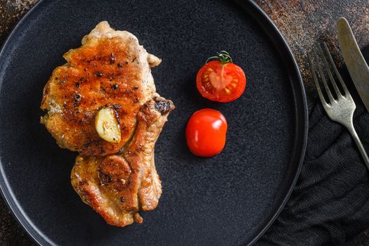 Grilled pork steak on the bone with seasonings on black dish with sfork and knife on cloth, over rustic metall surface top view close up.