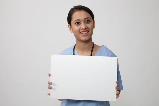 Nurse showing medical sign billboard standing, Young smiling woman nurse or doctor in scrubs showing empty blank sign board with copy space. Indian model isolated on white background