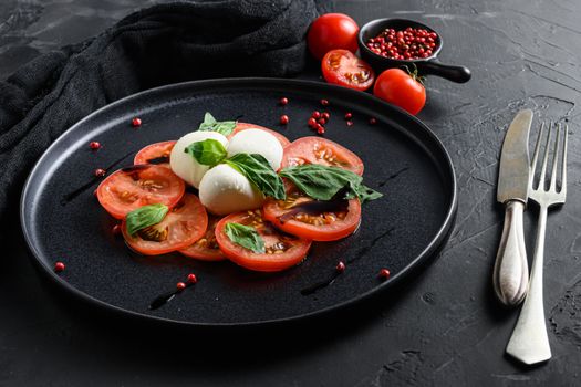Caprese salad Italian cuisine concept Tomato and mozzarella slices with basil leaves on black ceramic platwantipasta black textured background close up with fork and knife.