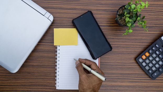 Writing on a notepad while working from home. A lap top, a mobile, calculator and plant are also on display on this brown striped working table.