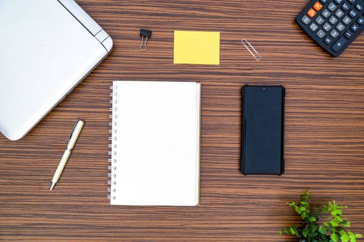 An office table working enviroment. Notepad, sticky note, pen plant, calculator and a lap top on a brown striped zebrawood design table top. Must have objects while working from home during Covid-19