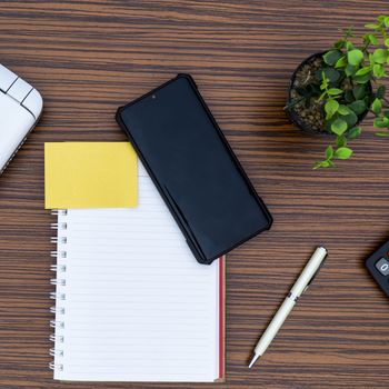 Notepad, sticky note, pen plant, calculator and a lap top on a brown striped zebrawood design table top. Must have objects while working from home during Covid-19