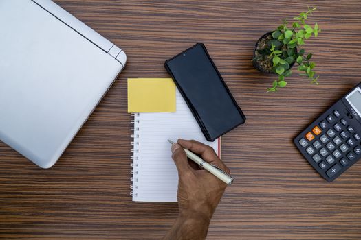 Writing on a notepad while working from home. A lap top, a mobile, calculator and plant are also on display on this brown striped working table.