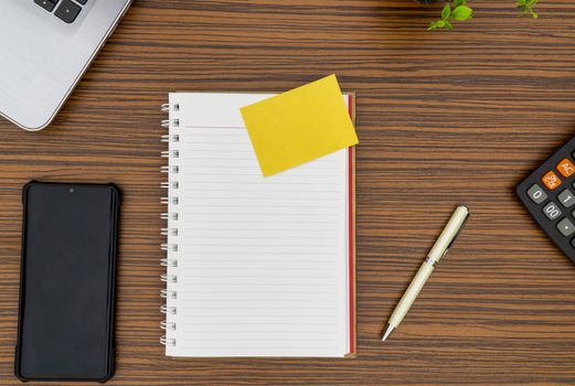 Notepad, sticky note, pen plant, calculator and a lap top on a brown striped zebrawood design table top. Must have objects while working from home during Covid-19