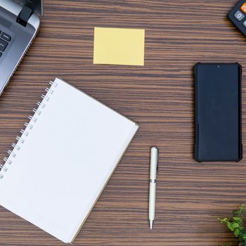 An office table working enviroment. Notepad, sticky note, pen plant, calculator and a lap top on a brown striped zebrawood design table top. Must have objects while working from home during Covid-19