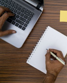 Writing on a notepad and typing on a keyboard while working on a project. A lap top, a mobile, calculator and plant are also on display on this brown striped working table.