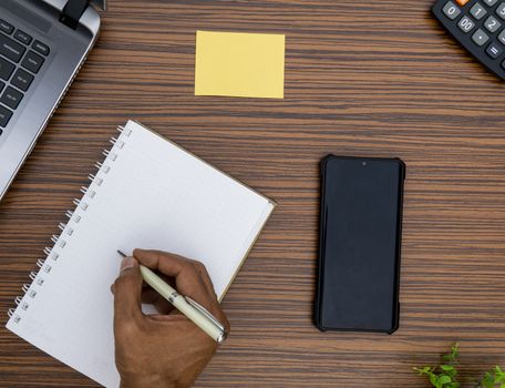 Writing on a notepad while working from home. A lap top, a mobile, calculator and plant are also on display on this brown striped working table.