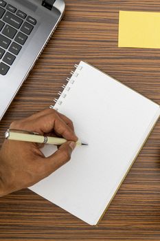 Writing on a notepad while working from home. A lap top, a mobile, calculator and plant are also on display on this brown striped working table.