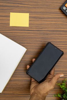 A brown hand holding a mobile phone while working in an office environment. A lap top, a mobile, calculator and plant are also on display on this brown striped working table.