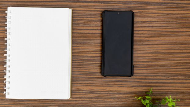 Personal note book and a black mobile phone with a glimpse of green plant on a striped brown office table