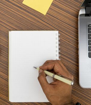 Writing on a notepad while working from home. A lap top, a mobile, calculator and plant are also on display on this brown striped working table.