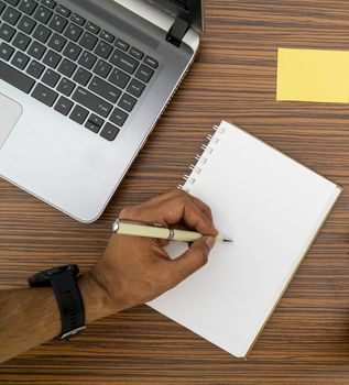 Writing on a notepad while working from home. A lap top, a mobile, calculator and plant are also on display on this brown striped working table.