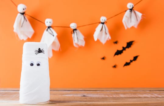 Funny Halloween day decor party concept, The mummy ghost on water glass wrapped around with bandage and have bats and spider stick it found only eyes, studio shot isolated on orange background