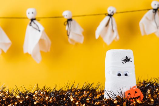 Funny Halloween day decor party concept, The mummy ghost on water glass wrapped around with bandage and have bats and spider stick it found only eyes, studio shot isolated on yellow background