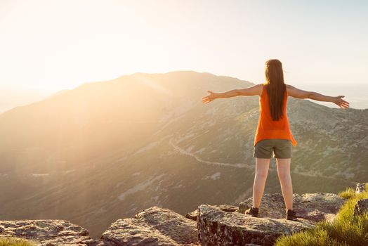 Happy woman with open arms stay on the peak of the mountain cliff edge under sunset light