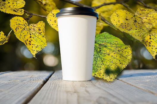 Paper cup with coffee is standing on the wooden table in on the autumn park with yellow leaves background. Craft cup of coffee on the road.