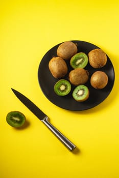 Kiwi fruits half sliced in black plate with kitchen knife on yellow background, top view