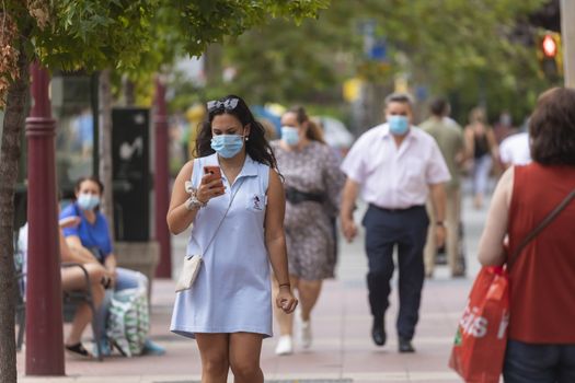 Zaragoza, Spain - August 18, 2020: A woman browse smartphone, while walks with face mask, due to the coronavirus pandemic, in the central area of Zaragoza.