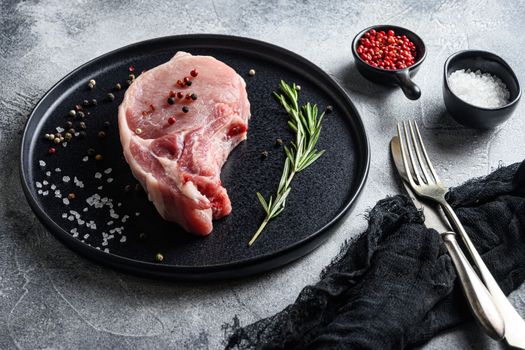 Raw Pork Loin chops in a black round plate on a grey textured stone background with rosemary garlic peppercorns ingredients for grill side view